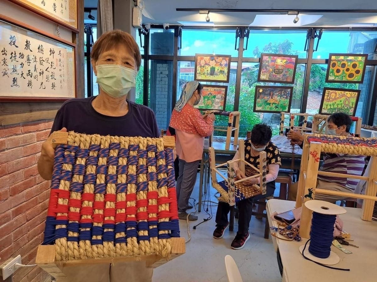長輩展示親手編織的椅凳坐墊，後方夥伴正專心編織